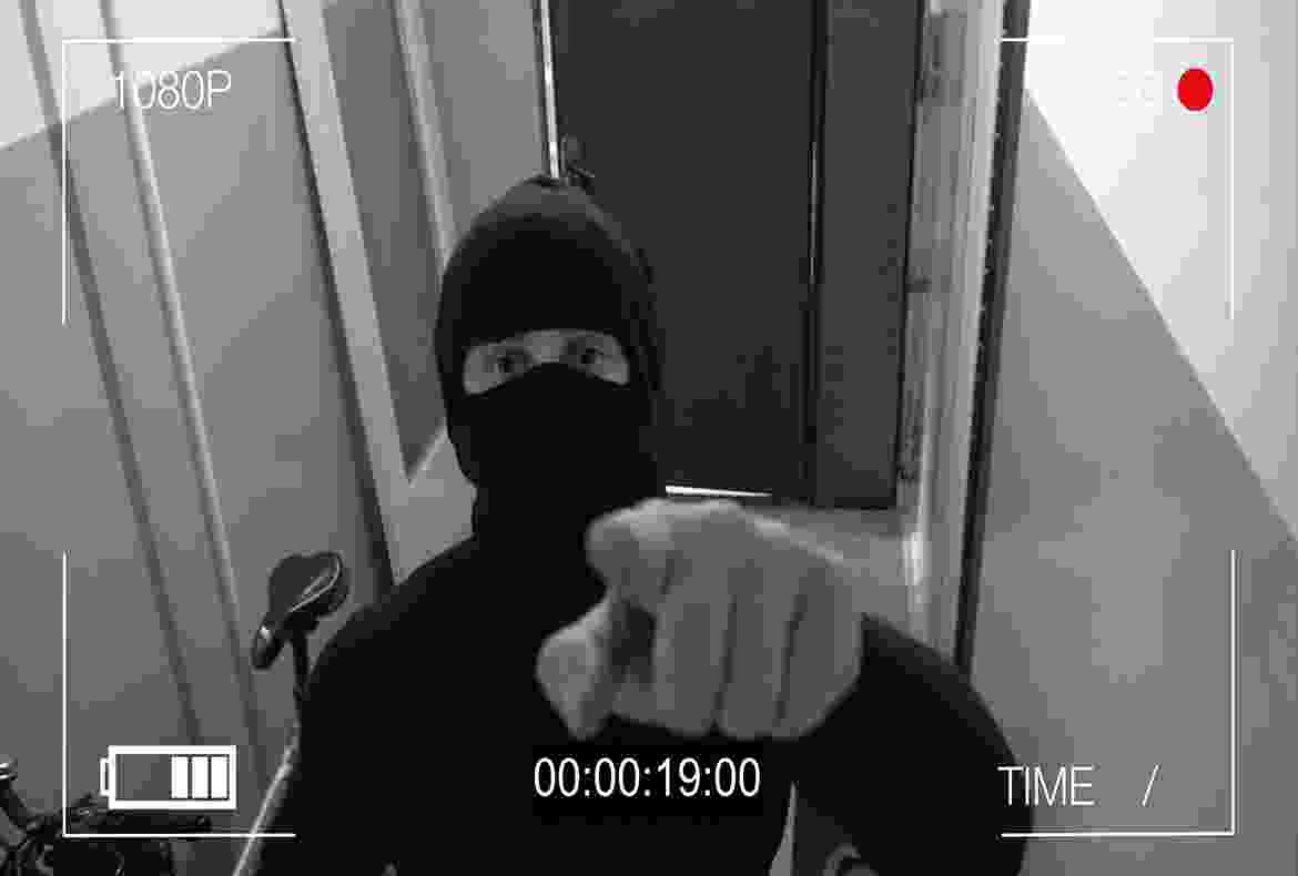 A person in a black mask pointing at the camera in a dimly lit hallway.