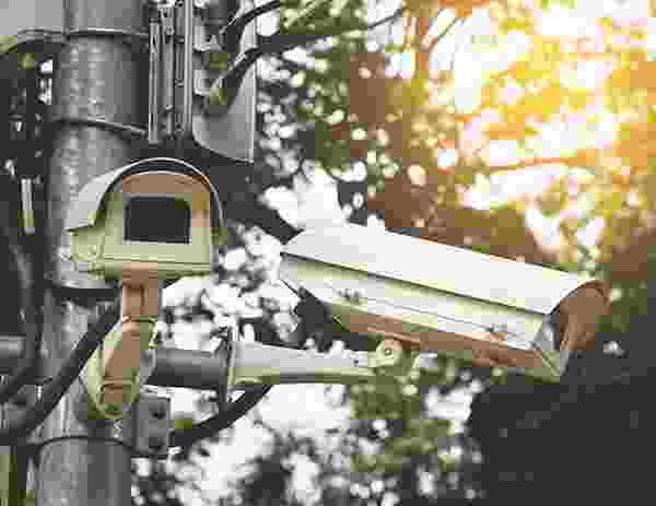 Two outdoor security cameras mounted on a pole with sunlight filtering through trees. Two outdoor security cameras mounted on a pole with sunlight filtering through trees.