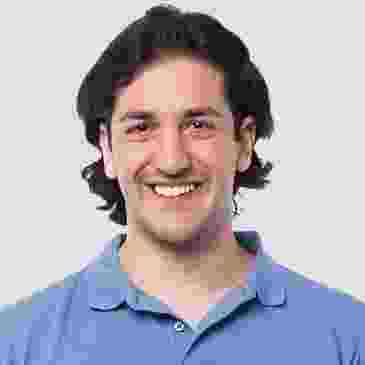 Smiling young man with dark hair in a blue shirt against a plain background.